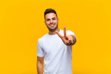 Genç İspanyol adam gülümsüyor ve mutlu görünüyor, kaygısız ve pozitif, turuncu duvara karşı zafer ya da barış işareti yapıyor.