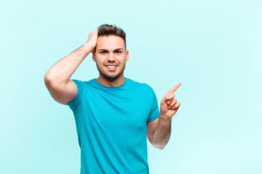 Genç İspanyol adam gülüyor, mutlu, pozitif ve şaşırmış görünüyor, mavi arkaplana karşı yanal fotokopi alanını işaret eden harika bir fikri fark ediyor.