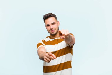 Genç İspanyol adam mutlu ve kendinden emin, kamerayı iki eliyle gösteriyor ve gülüyor, mavi duvara karşı seni seçiyor.