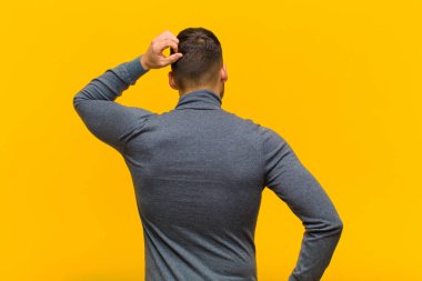 Genç İspanyol bir adam bilgisiz ve şaşkın hissediyor, bir çözüm düşünüyor, eli kalçasında, diğeri kafasında, turuncu duvara karşı dikiz manzaralı