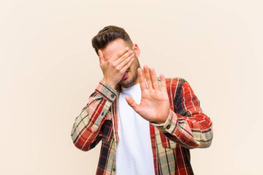 Latin asıllı genç bir adam elleriyle yüzünü örtüyor ve diğer elini kamerayı durdurmak için öne koyuyor. İzole edilmiş arka planda fotoğraf ve resimleri reddediyor.