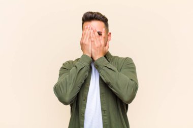 Genç İspanyol adam elleriyle yüzünü kapatıyor, parmaklarının arasından şaşkın bir ifadeyle bakıyor ve izole edilmiş arka plana bakıyor.