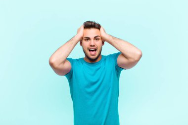 Genç İspanyol adam ellerini kaldırıyor, ağzı açık, son derece şanslı, şaşkın, heyecanlı ve mutlu, mavi arka planda