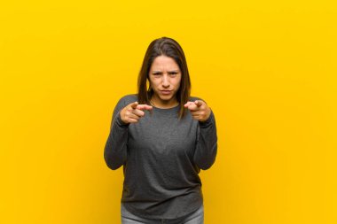 Latin Amerikalı kadın kameraya hem parmaklarıyla hem de kızgın bir ifadeyle bakıyor ve sana sarı duvara karşı görevini yapmanı söylüyor.