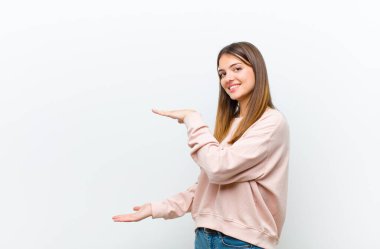 Genç, güzel bir kadın gülümsüyor, mutlu, pozitif ve tatmin olmuş hissediyor, beyaz arkaplana karşı fotokopi alanında nesne ya da konsept tutuyor ya da gösteriyor