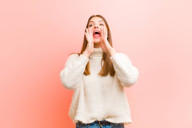 Genç, güzel bir kadın mutlu, heyecanlı ve pozitif hissediyor. Elleri ağızda, pembe arka planda bağırıyor.