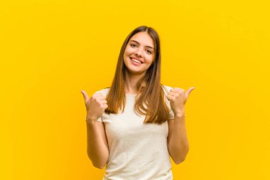 Genç, güzel bir kadın neşeyle gülümsüyor ve mutlu görünüyor. Kaygısız ve pozitif hissediyor. İki başparmağı da turuncu arka planda.