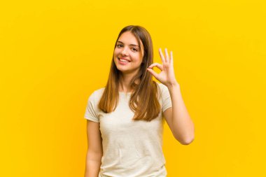 Genç, güzel bir kadın mutlu, rahat ve tatmin olmuş hissediyor. Onayını iyi bir jestle gösteriyor. Turuncu arka planda gülümsüyor.