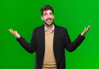 Genç işadamı kendini mutlu, şaşkın, şanslı ve şaşırmış hissediyor. İki eli havada, yeşil arka planda zaferi kutluyor.
