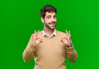 Genç işadamı şaşkın, şaşkın ve şaşırmış hissediyor, yeşil arka plana karşı iki elle onay işareti yapıyor.