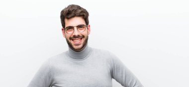 Genç yönetici mutlu bir şekilde gülümsüyor, eli kalçasında ve kendinden emin, pozitif, gururlu ve beyaz duvara karşı dost canlısı bir tavırla.