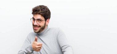 Genç yönetici kendini mutlu, sakin, tatmin olmuş, rahat ve başarılı hissediyor, kameraya bakıyor, beyaz duvara karşı seni seçiyor.