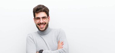 Genç yönetici kollarını kavuşturup mutlu bir şekilde gülüyor. Beyaz duvara karşı rahat, olumlu ve tatmin edici bir poz veriyor.