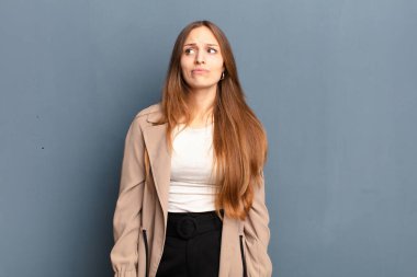 Genç ve güzel bir kadın şaşkın ve şaşkın görünüyor, merak ediyor ya da bir sorunu çözmeye çalışıyor ya da gri arka plana karşı düşünüyor.