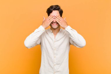 Genç yakışıklı adam gülümsüyor ve mutlu hissediyor, iki eliyle gözlerini kapatıyor ve turuncu arka planda inanılmaz bir sürpriz bekliyor.