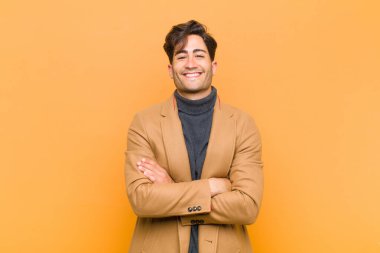 Genç yakışıklı adam mutlu, gururlu ve tatmin olmuş görünüyor. Kolları turuncu arka planda birleşmiş başarılı bir şekilde gülümsüyor.