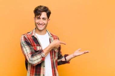 Genç yakışıklı adam neşeyle gülümsüyor ve yan taraftaki fotokopi alanını işaret ederek turuncu arka planda bir nesneyi gösteriyor ya da reklamını yapıyor.