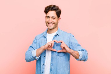 Genç yakışıklı adam gülümsüyor ve mutlu, sevimli, romantik ve aşık, pembe arka planda iki eliyle kalbi şekillendiriyor.