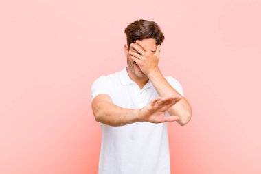 Genç ve yakışıklı bir adam elini yüzünü örtüyor ve diğer elini kamerayı durdurmak için öne koyuyor. Pembe arka plandaki fotoğraf ve resimleri reddediyor.