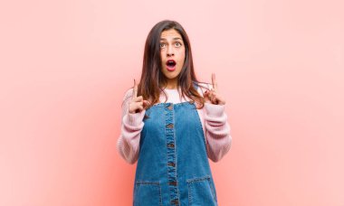 Genç, güzel bir kadın şaşkın, şaşkın ve ağzı açık görünüyor. Pembe arka planda uzayı kopyalamak için iki elini havaya kaldırıyor..