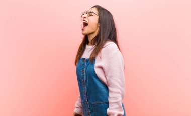 Genç, güzel bir kadın öfkeyle bağırıyor, agresif bir şekilde bağırıyor, pembe arka planda stresli ve kızgın görünüyor..