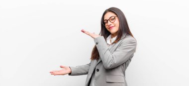 Genç ve güzel bir kadın, iki eli de kenarda olan bir objeyi tutuyor. Kopya boşluğunda, beyaz duvara yaslanmış bir objeyi gösteriyor, sunuyor ya da reklamını yapıyor.