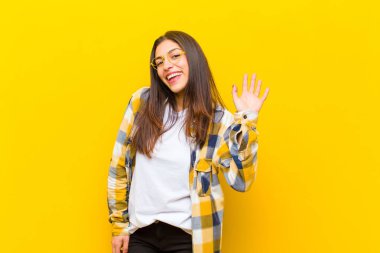 Genç ve güzel bir kadın mutlu bir şekilde gülümsüyor, el sallıyor, seni selamlıyor, ya da turuncu duvara veda ediyor.