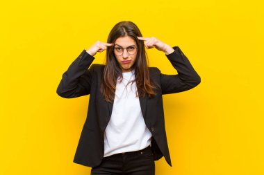 Ciddi ve konsantre bakışları olan genç ve güzel bir kadın, turuncu duvara karşı zorlu bir sorun düşünüyor ve beyin fırtınası yapıyor.