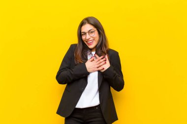 Genç, güzel bir kadın romantik, mutlu ve aşık, neşeli bir şekilde gülümsüyor ve turuncu duvara karşı kalbe yakın el ele tutuşuyor.