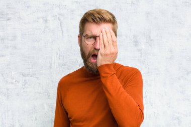 Uykulu görünüyor, sıkılmış ve esniyor, baş ağrısı ve yüzünün yarısını kaplayan bir elle.