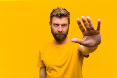 Genç sarışın adam ciddi, sert, hoşnutsuz ve kızgın görünüyor. Avuç içini açıp turuncu duvara karşı jest yapıyor.