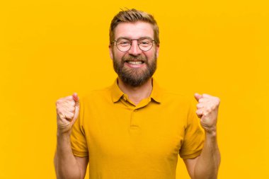 Genç sarışın adam zaferle bağırıyor, gülüyor, mutlu ve heyecanlı turuncu duvara karşı başarıyı kutlarken.