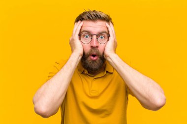 Genç sarışın adam hoş olmayan bir şekilde şok olmuş, korkmuş ya da endişeli görünüyor, ağzı sonuna kadar açık ve turuncu duvara karşı iki kulağını da kapatıyor.