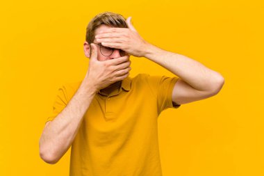 Genç sarışın adam kameraya hayır diyen iki eliyle yüzünü kapatıyor! Resimleri reddetmek ya da turuncu duvara karşı fotoğraf yasaklamak