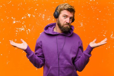 Genç sarışın adam dans ediyor ve müzik dinliyor. Kulaklıkları mor kapüşonlu. Zarar görmüş turuncu duvara karşı.