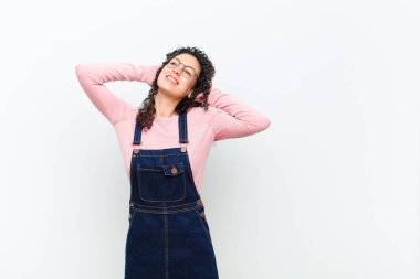 Genç ve güzel bir kadın gülümsüyor ve rahatlıyor, tatmin oluyor ve kaygısız, pozitif gülüyor ve beyaz duvara yaslanıyor.