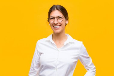 Genç iş kadını mutlu bir şekilde gülümsüyor, eli kalçasında ve kendinden emin, pozitif, gururlu ve turuncu duvara karşı dost canlısı bir tavırla.