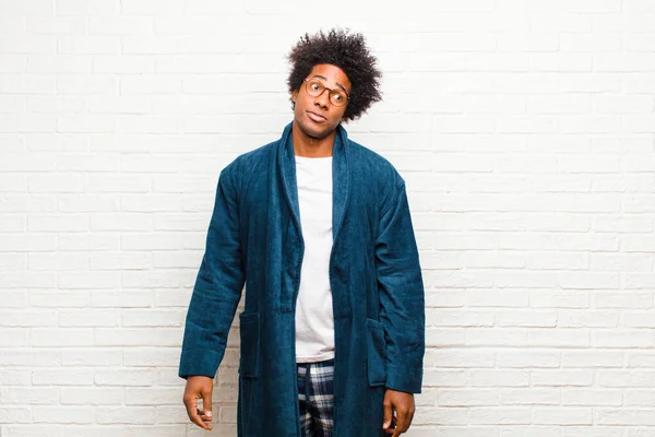 young black man wearing pajamas with gown wondering, thinking happy thoughts and ideas, daydreaming, looking to copy space on side against brick wall