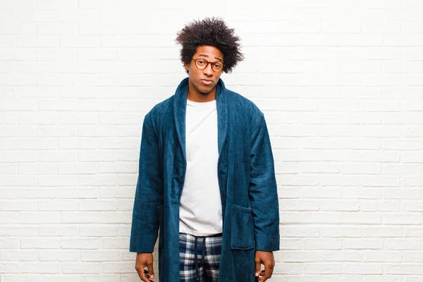 young black man wearing pajamas with gown feeling sad and whiney with an unhappy look, crying with a negative and frustrated attitude against brick wall