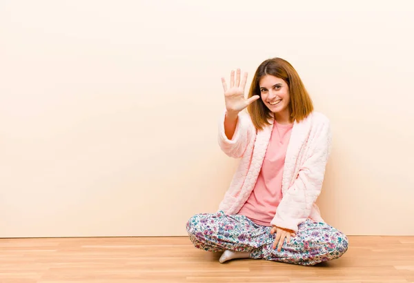 young woman wearing pajamas sitting at home smiling and looking friendly, showing number five or fifth with hand forward, counting down