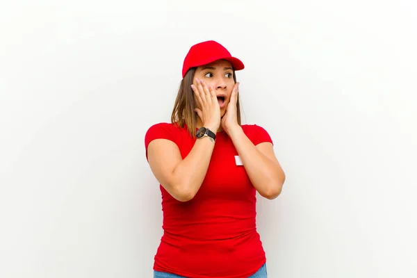 delivery woman feeling happy, excited and surprised, looking to the side with both hands on face against white background