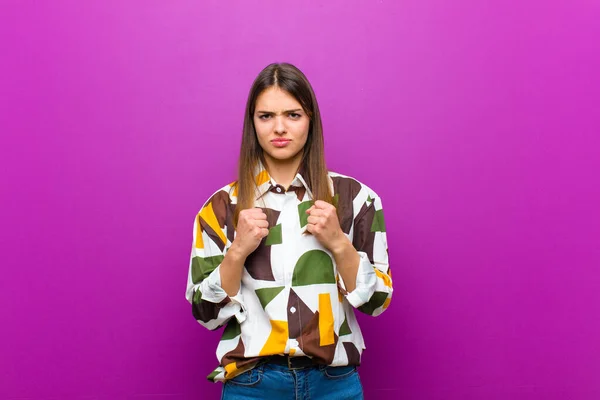 young pretty woman looking confident, angry, strong and aggressive, with fists ready to fight in boxing position against purple background