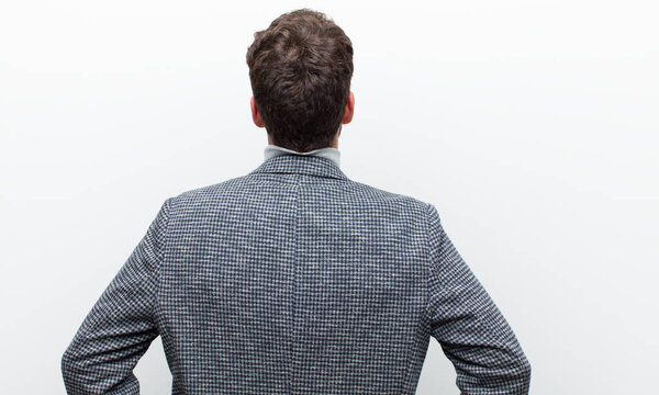 young manager man feeling confused or full or doubts and questions, wondering, with hands on hips, rear view against white wall