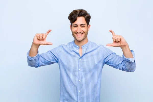 young handsome man framing or outlining own smile with both hands, looking positive and happy, wellness concept against blue background
