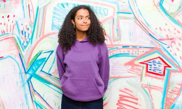 young pretty african american woman looking puzzled and confused, wondering or trying to solve a problem or thinking against graffiti wall