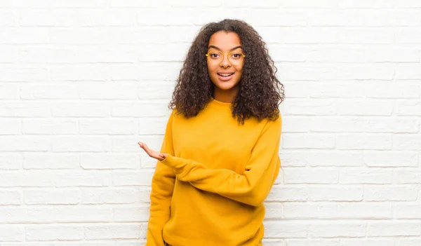 young black woman smiling cheerfully, feeling happy and showing a concept in copy space with palm of hand against brick wall