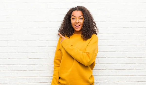 young black woman looking excited and surprised pointing to the side and upwards to copy space against brick wall