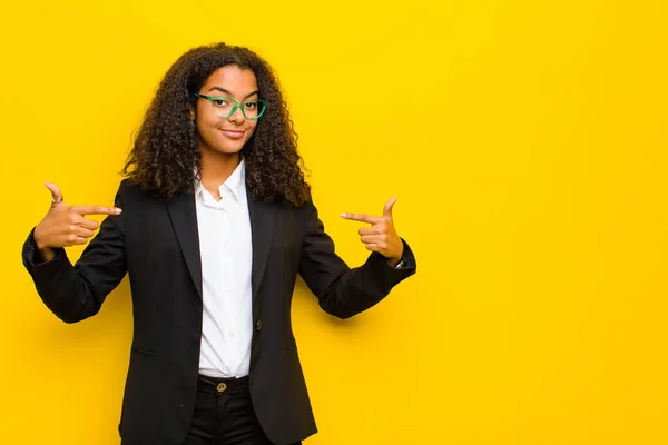 black business woman looking proud, arrogant, happy, surprised and satisfied, pointing to self, feeling like a winner against orange wall