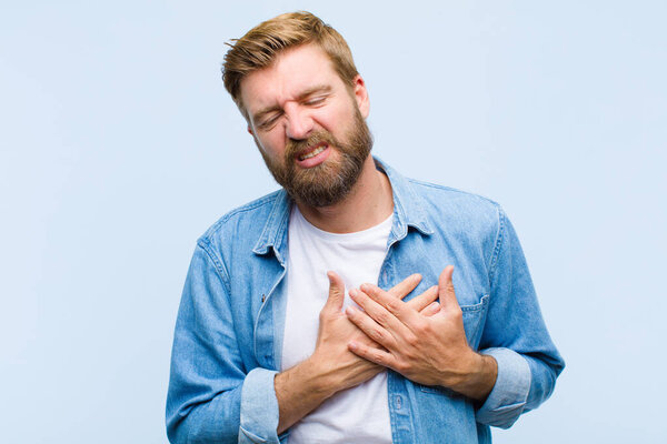 young blonde adult man looking sad, hurt and heartbroken, holding both hands close to heart, crying and feeling depressed