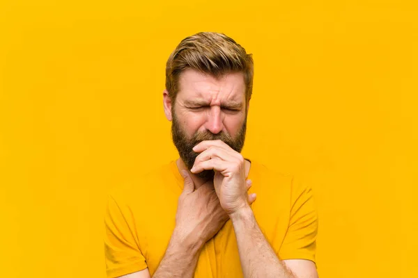young blonde man feeling ill with a sore throat and flu symptoms, coughing with mouth covered against orange wall